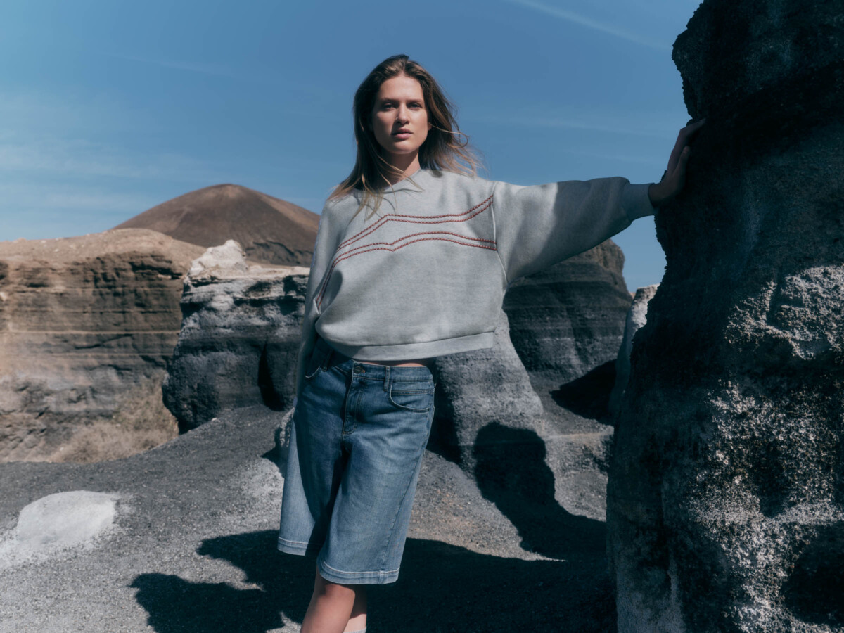 Lanius: Frau, die in einer steinigen Naturlandschaft steht, bekleidet mit einem grauen Baumwollpullover mit Rundhalsausschnitt und roter Stickerei sowie blauen Jeansshorts.