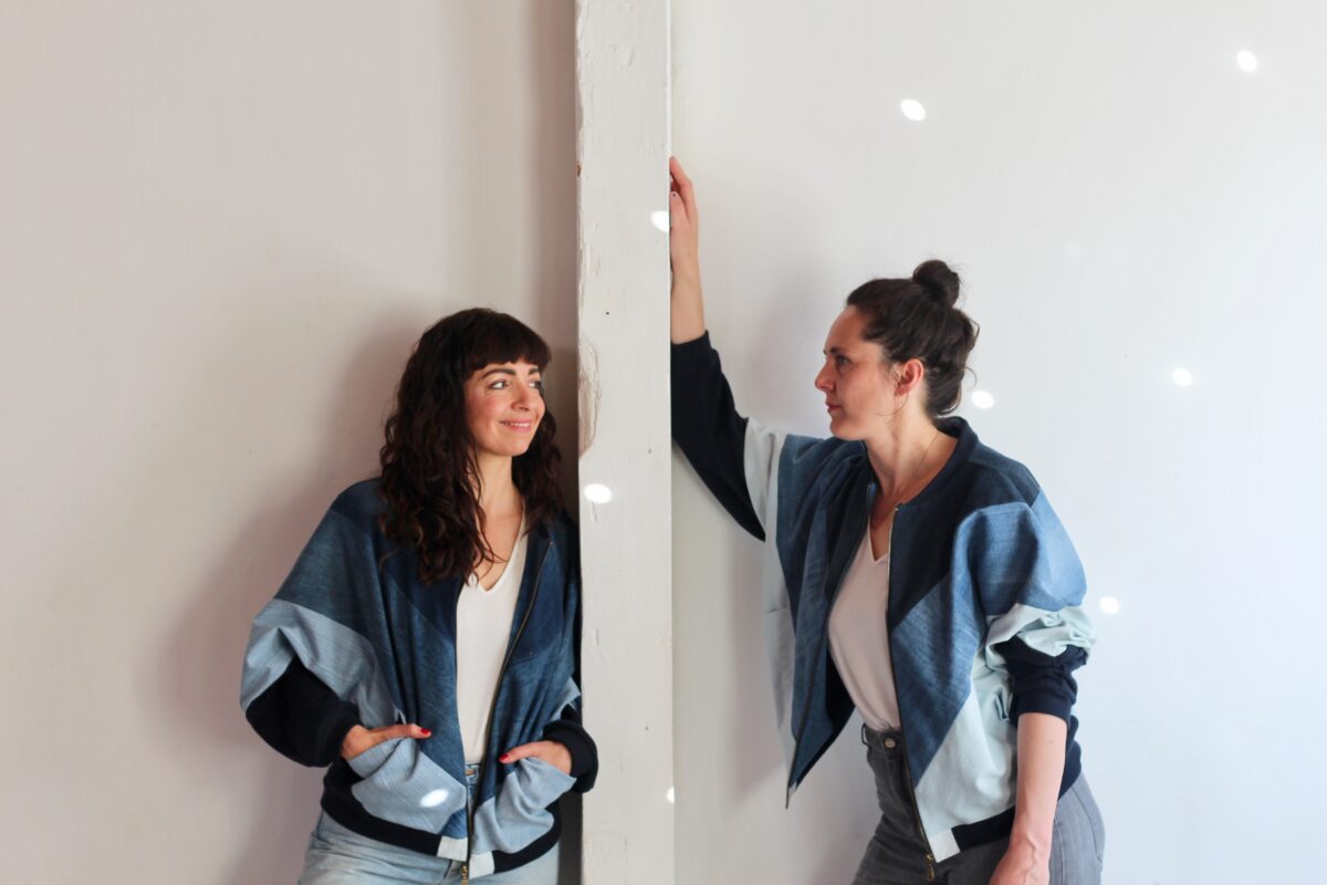 Die Gründerinnen von Bridge & Tunnel, Conny und Lotte, lehnen an einer Wand und schauen sich an. Beide tragen einen Blouson aus alten Jeansstoffen