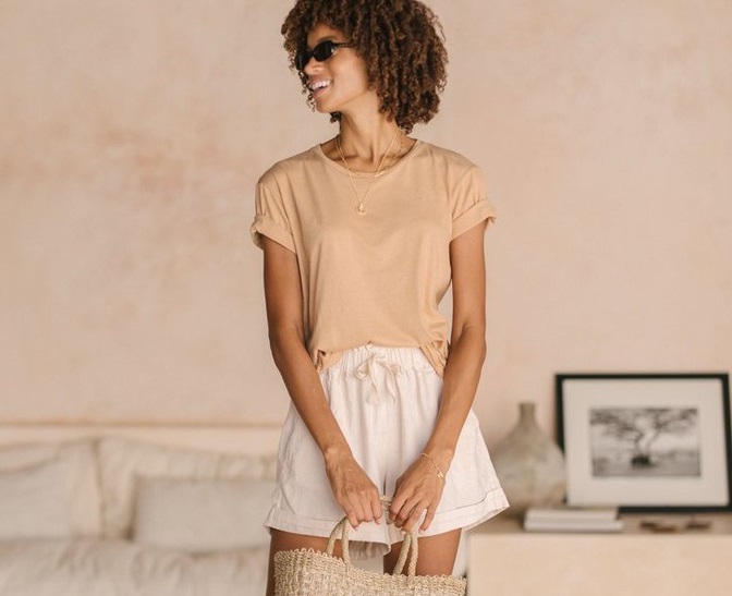 Frauen trägt helles T-Shirt und weiße Shorts und hält eine Basttasche vor sich