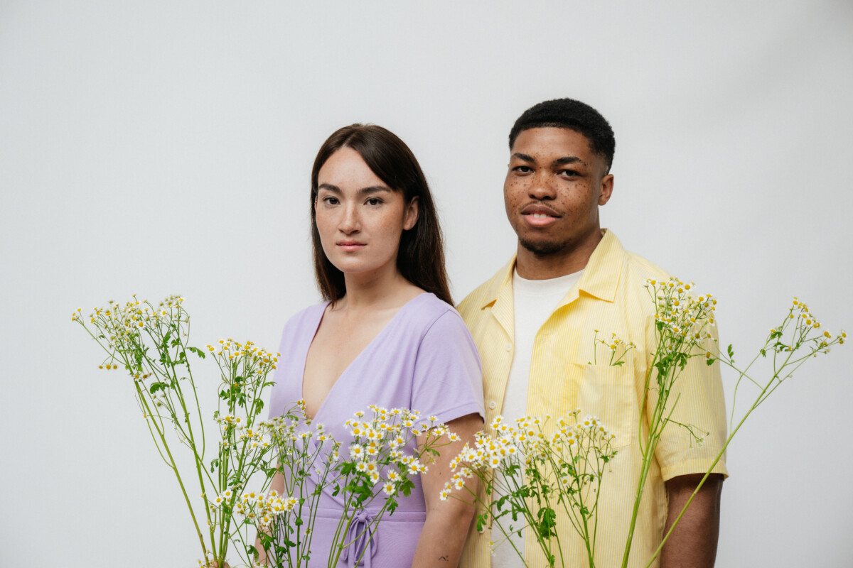 MELA Wear - ein weibliches und ein männliches Model stehen hinter einem Strauch Blumen. Das weibliche Model trägt ein fliedernes kurzärmliges Kleid mit einem tiefen V-Ausschnitt, das männliche Model trägt ein pastellgelbes kurzärmliges