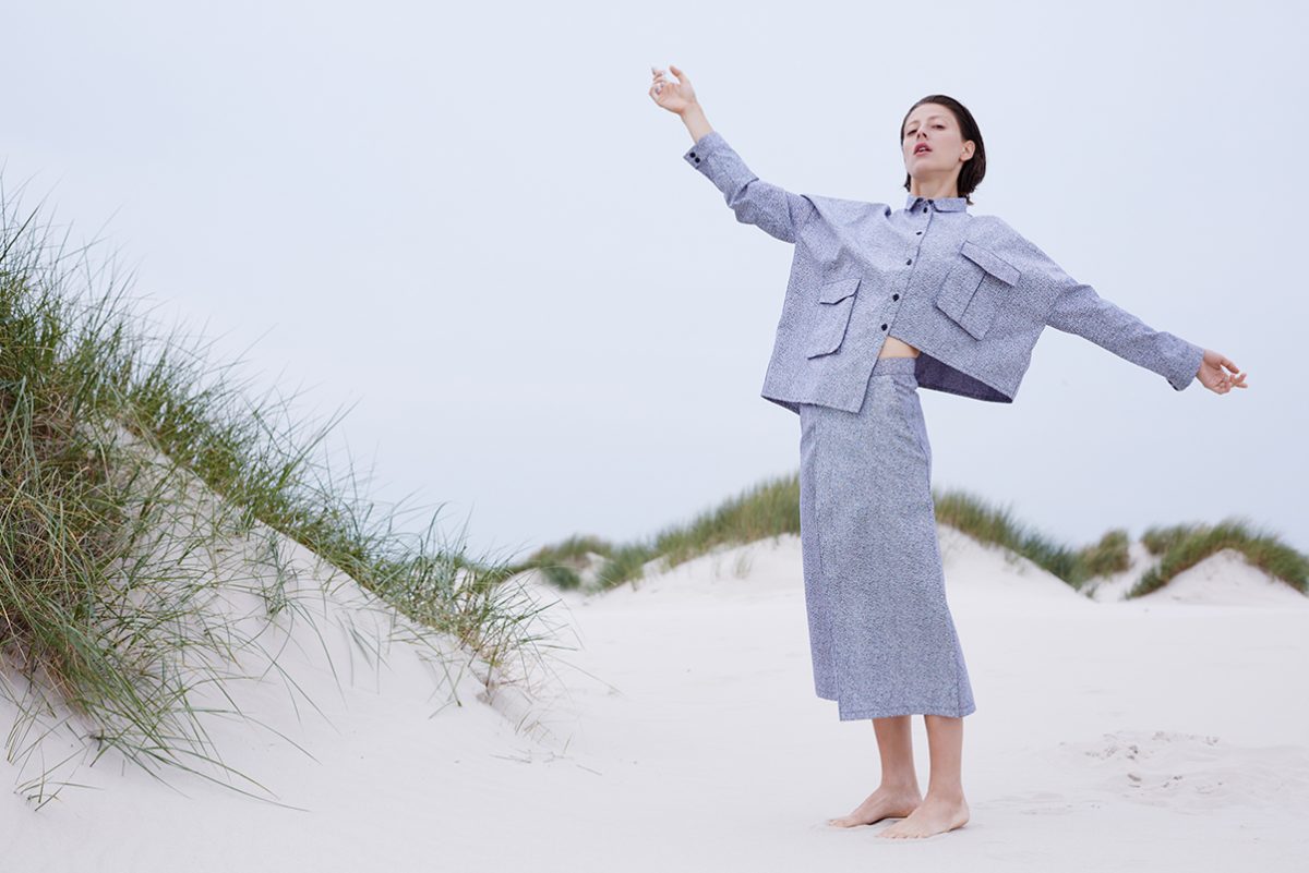 Frau im grauen Zweiteiler mit ausgebreiteten Armen auf einer Sanddüne