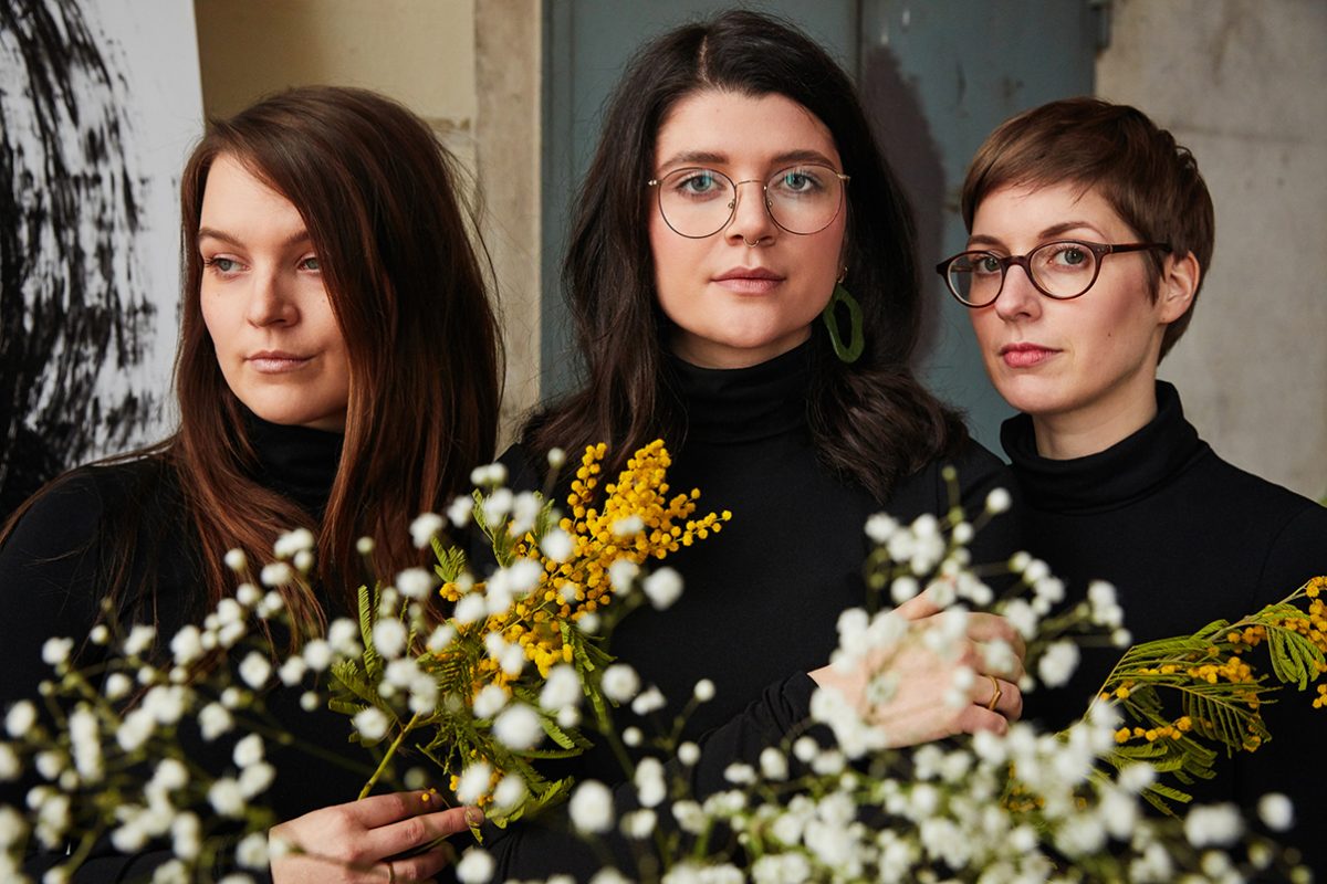 3 Frauen tragen schwarze Rollkragenpullover, alle halten weiß-gelbe Blumen