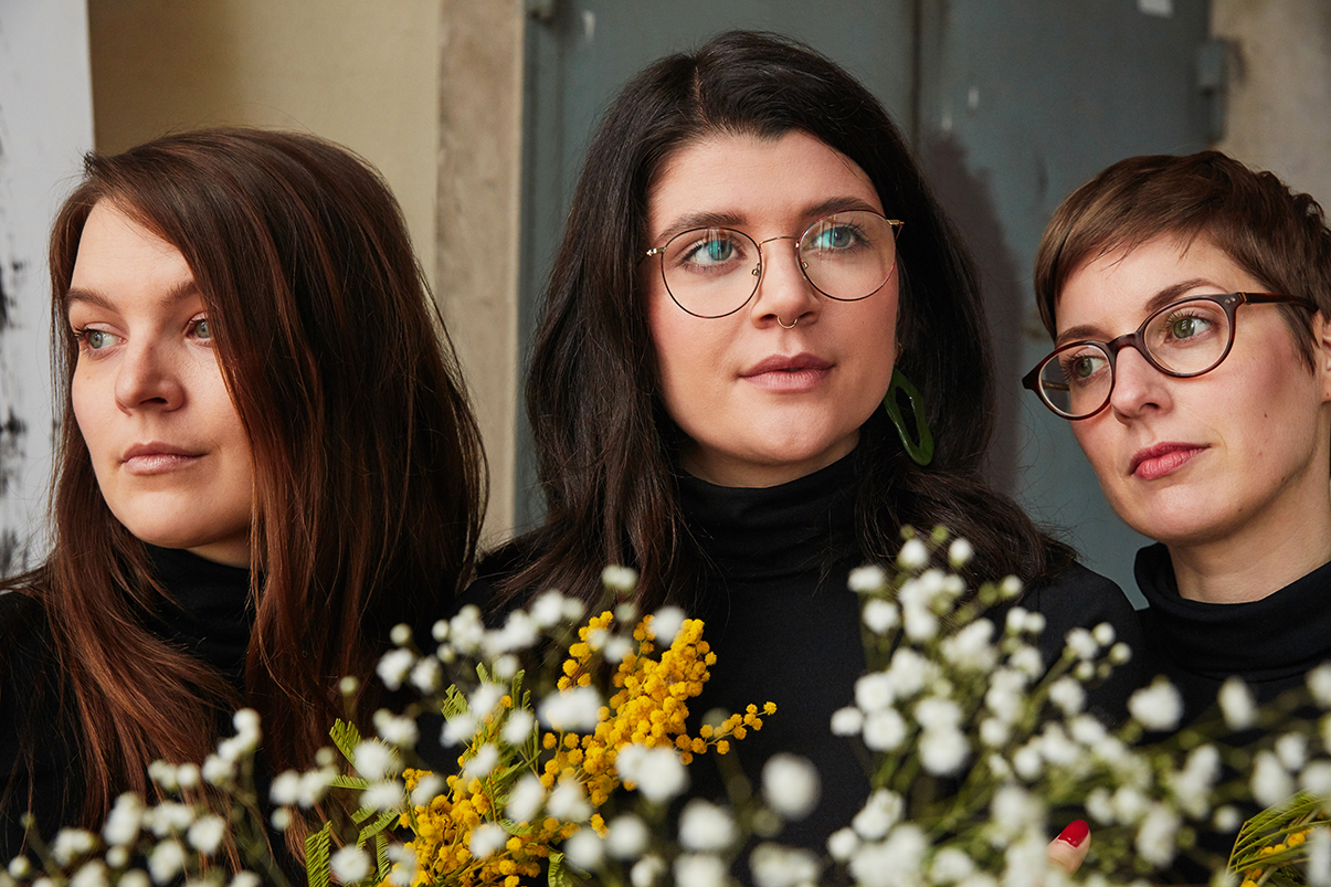 3 Frauen mit schwarzen Rolkragenpullovern von Wolford stehen eng beinander und schauen in unterschiedliche Richtungen, im Bildvordergrund sind weiß-gelbe Blumen