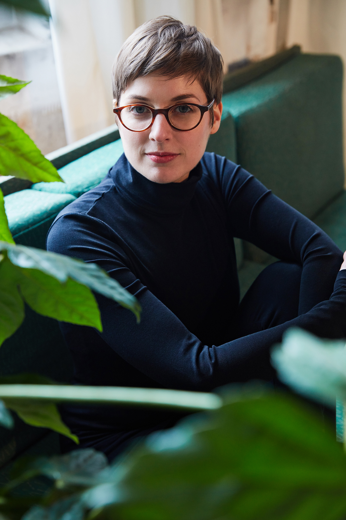 Frau im schwarzen Rollkragenpullover und schwarzer Leggings von Wolford sitzt mit angezogenne Knien auf einem grünen Sofa