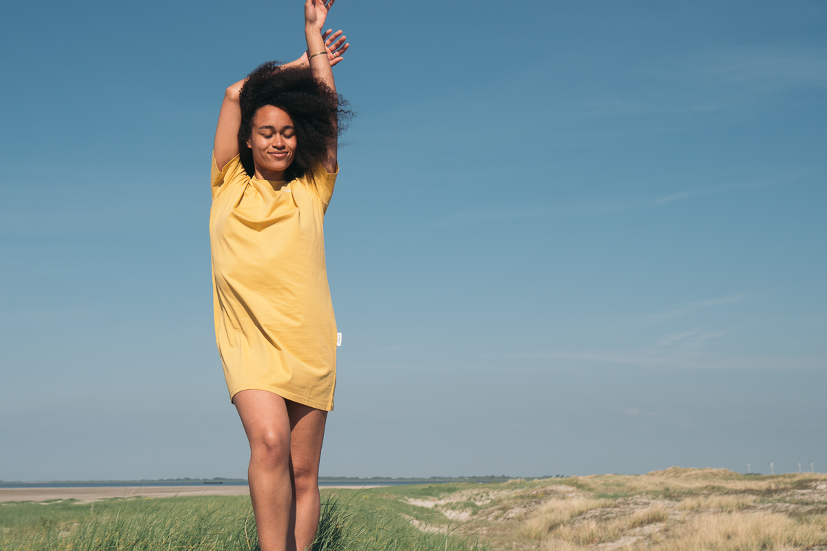 Ein weiblich gelesenes Model steht vor einer naturbelassenen Landschaft mit blauem Himmel. Sie trägt ein sonnengelbes T-Shirt Kleid. Ihre Arme streckt sie in die Höhe und hat dabei ihre Augen geschlossen, als würde sie eine Brise genießen.