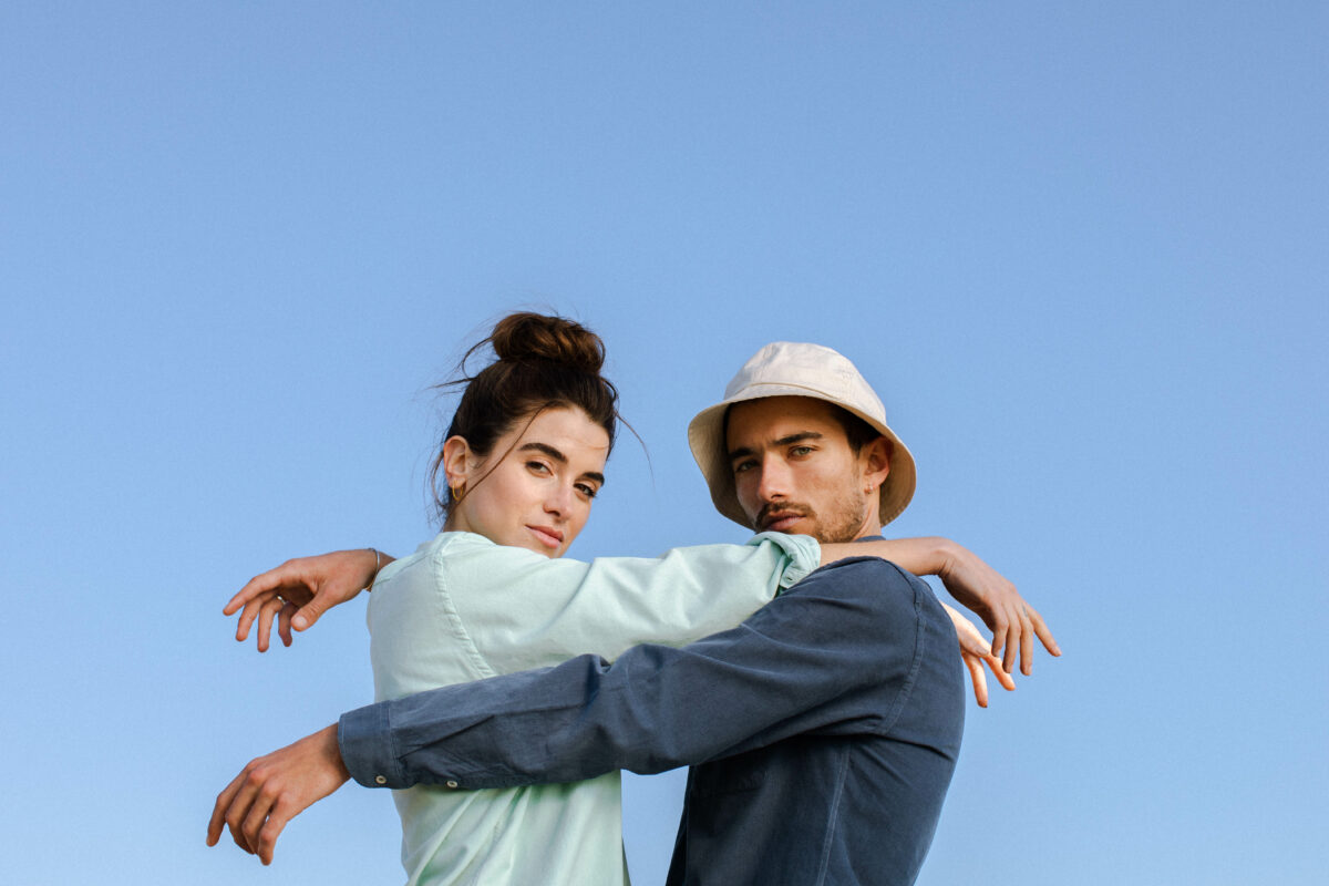 Shop like you give a damn, Vor blauem Himmel sind eine Mann mit Bucket Hat und eine Frau mit dunklen Haaren in einer statischen Umarmung, beide tragen ein Hemd, Womenswear,Menswear