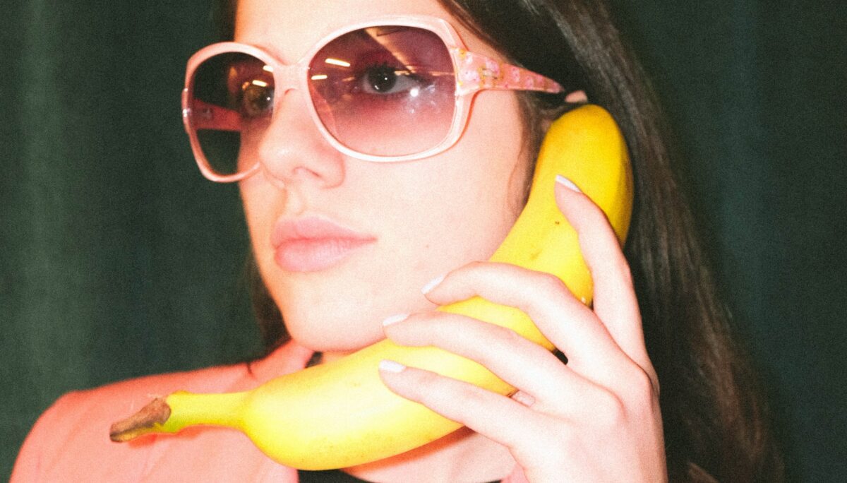 Model trägt eine große pinke Sonnenbrille, einen pinken Blazer und hält eine gelbe reife Banane als Telefon in der Hand