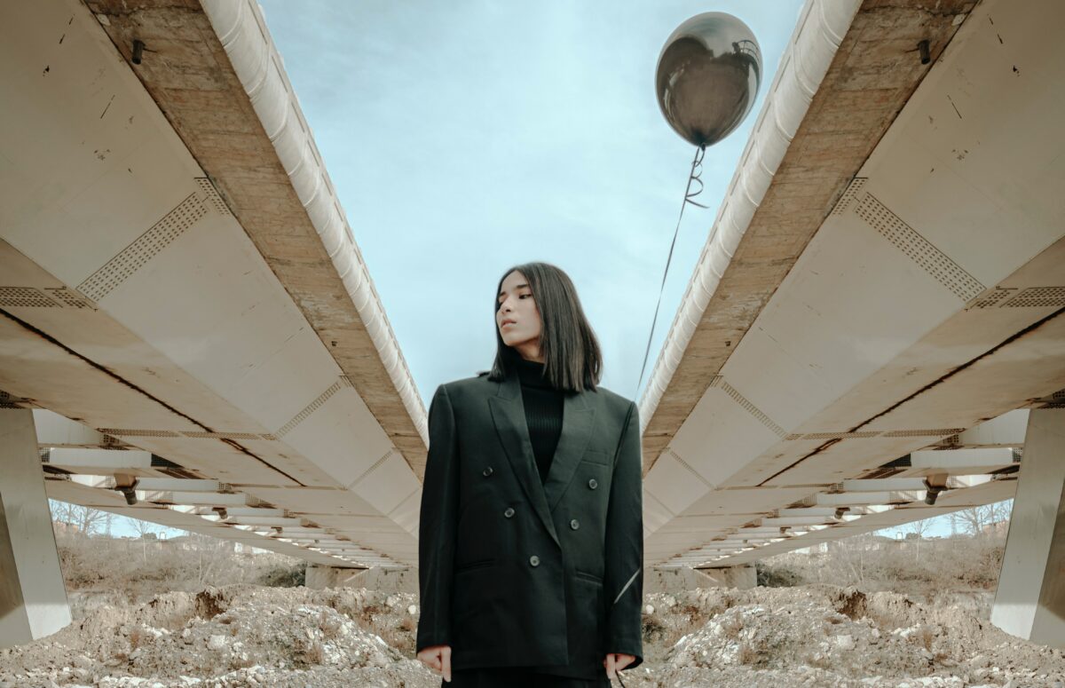Model trägt einen schwarzen Blazer, schwarzes Top und schwarze Hose, hält einen schwarzen Luftballon in den Händen und steht unter einer Brücke