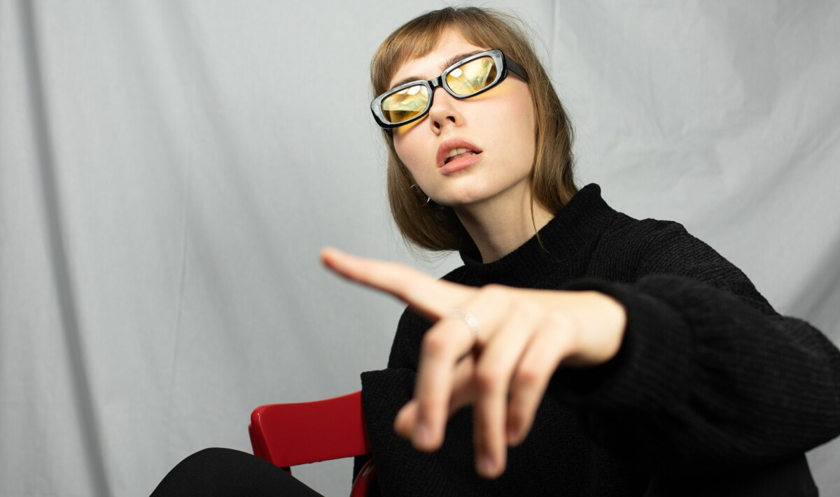 Greenwashing erkennen: Model sitzt auf einem roten Stuhl, trägt einen schwarzen Pullover, schwarze Sonnenbrille und zeigt mit dem Zeigefinger auf die Kamera