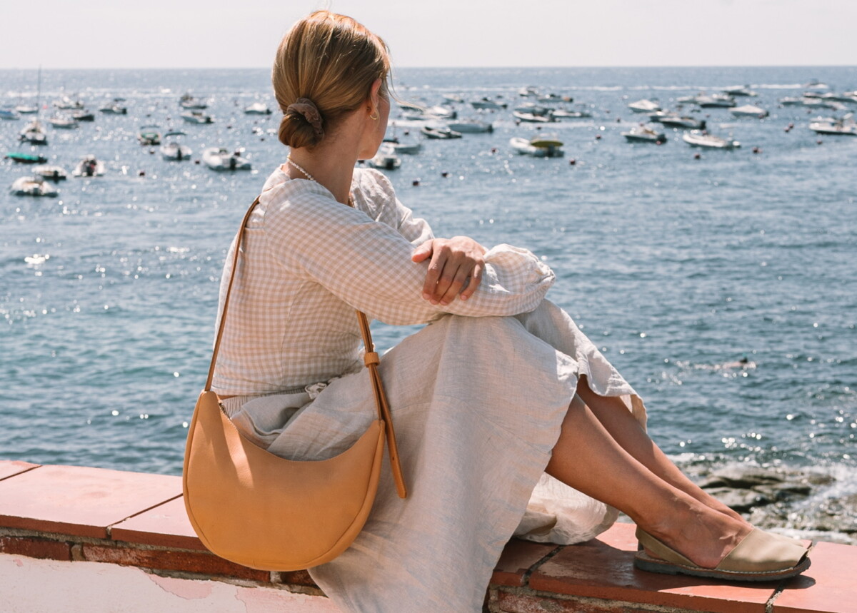 marin et marine: Eine Frau sitzt mit Blick auf das Meer, trägt eine nachhaltig gefertigte, karamellfarbene Lederhandtasche und ein weißes Baumwollkleid.