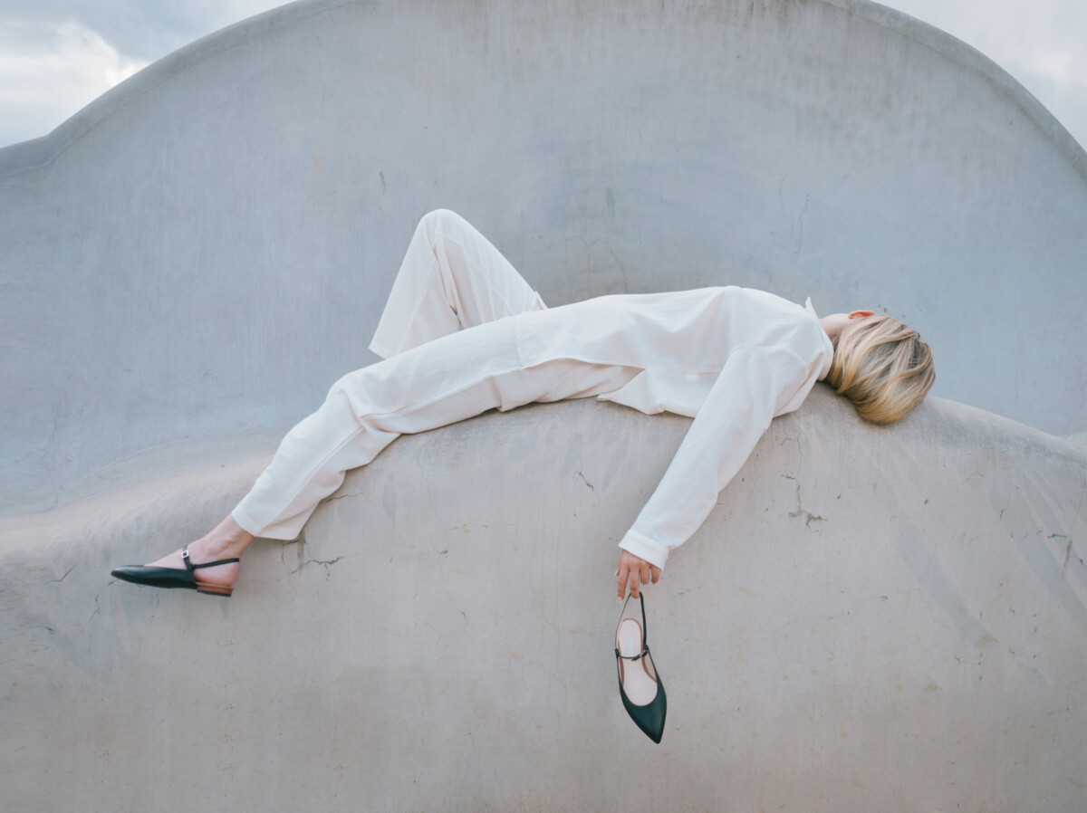 Corpado, Auf einer futuristischen Architektur aus hellem grau liegt eine Frau in weißem Anzug und hält einen Schuh in der linken Hand, Shoes