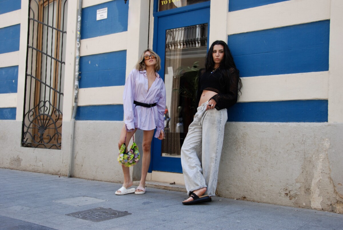 seyes shoes. Zwei Models stehen vor einer Hausfassage mit blauen und weißen Streifen. Das linke Model mit einem blonden Bob trägt eine Sonnenbrille, ein oversized fliederfarbiges Hemd, das mit einem Gürtel tailliert ist. Dazu hält sie eine bunte Tasche. Das rechte Modelträgt ihre dunklen welligen Haare offen, dazu ein gecropptes schwarzes Mesch-Oberteil und lockere helle Jeans. Beide Models tragen Schlappen, links in weiß und rechts in schwarz.