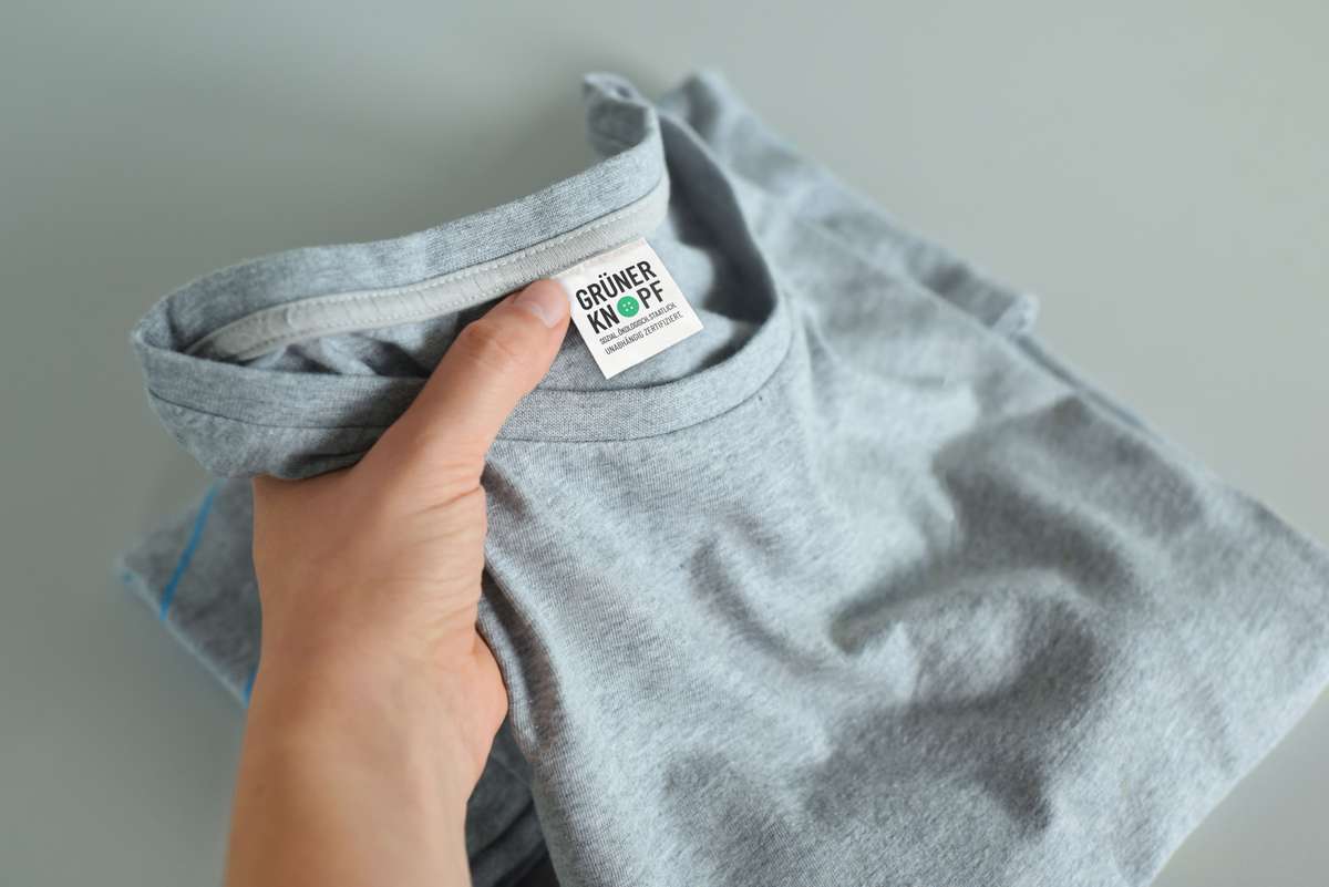 Graues T-Shirt mit Etikett, auf dem ein Grüner Knopf-Logo abgebildet ist