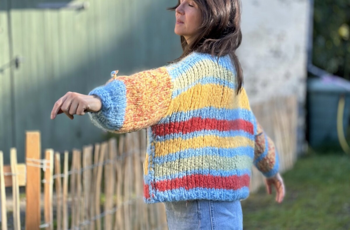 ViolasWollWerk: Eine Frau in einem bunten, grob gestrickten, übergroßen gestreiften Pullover schwingt draußen ihre Arme.